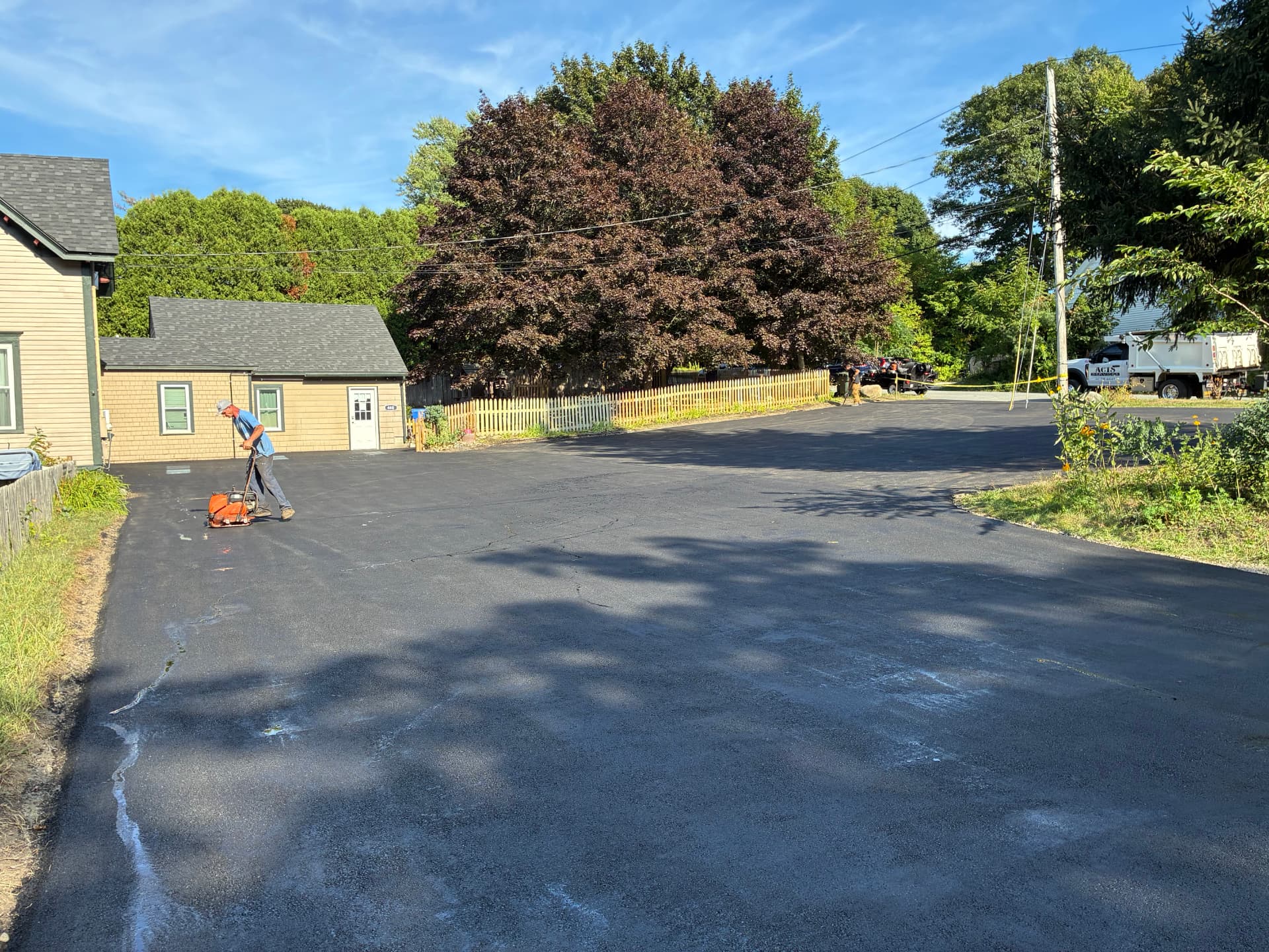ACTS Services crew member running a plate compactor along the edge of freshly laid asphalt with the company dump truck parked nearby