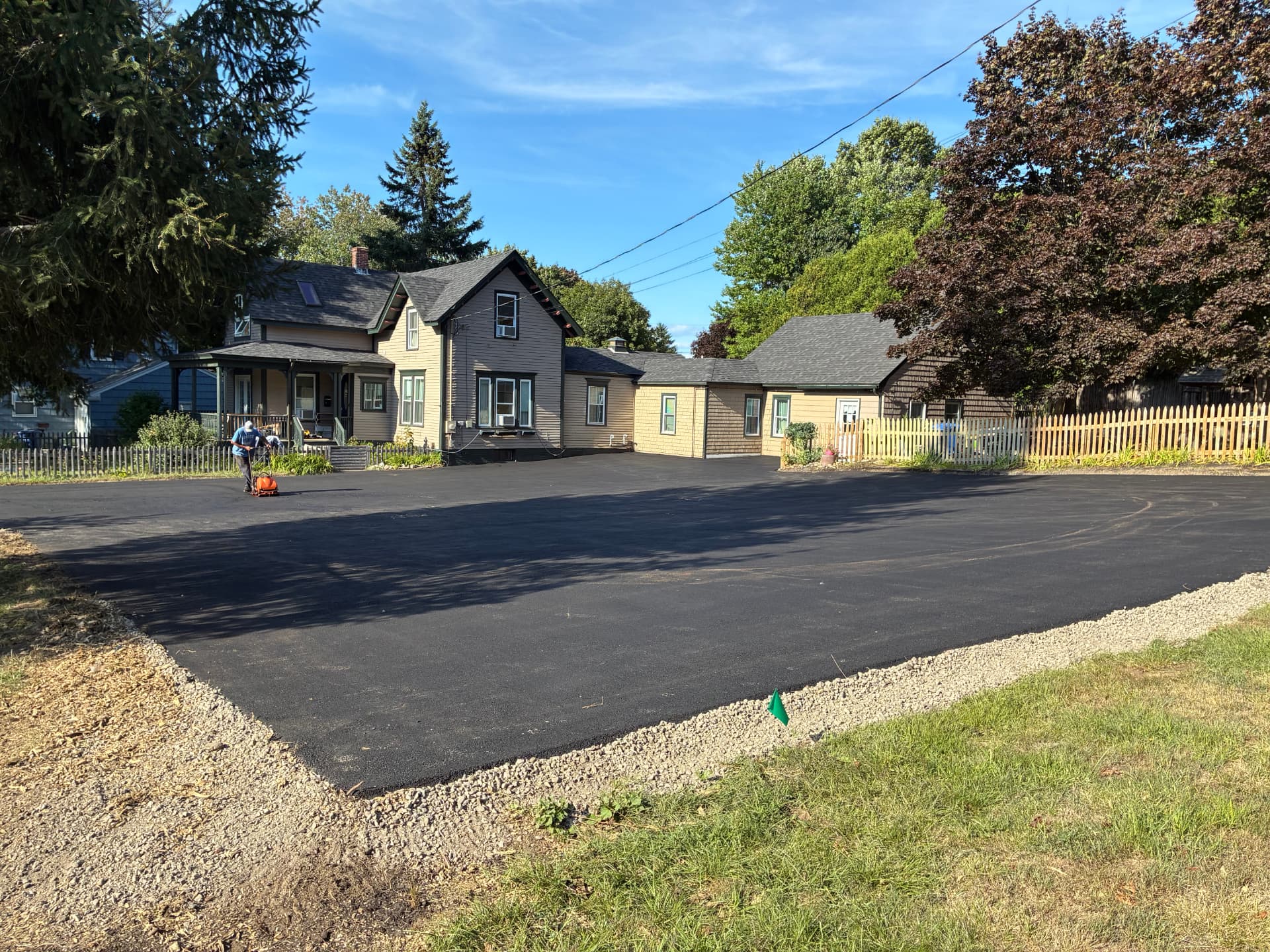 Freshly paved black asphalt parking area behind a tan farmhouse with a gravel edge shoulder in southern Maine