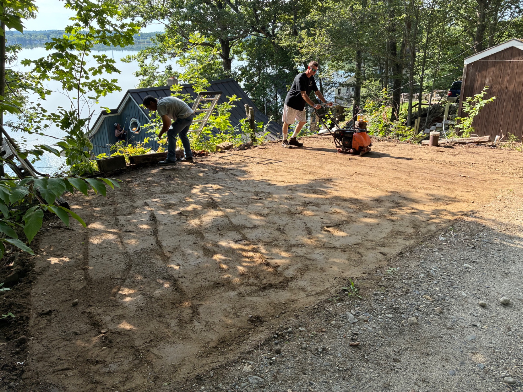 ACTS Services crew grading and compacting a gravel base for a parking pad on a wooded waterfront lot in Maine